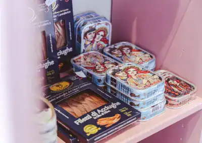 In einem Regal stehen Dosen mit Sardellen, auf deren Deckeln bunte Abbildungen von Frauen zu sehen sind, sowie rechteckige dunkle Schachteln mit der Aufschrift "Filetti di Acciughe". Die Dosen und Schachteln sind fein säuberlich auf einem rosa Regal angeordnet.