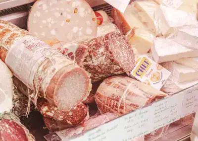 Verschiedene Wurst- und Käsesorten sind in einer Feinkostvitrine ausgestellt. Zu sehen sind verpackte Salami, Mortadella, Käseblöcke und eine Packung mit der Aufschrift "CHEF Parmigiano". Im Vordergrund befinden sich weiße Etiketten mit Text.