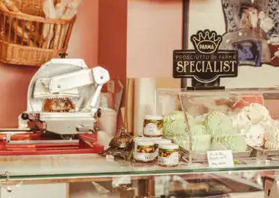 An der Feinkosttheke stehen Gläser mit Soßen, ein Schild mit Parmaschinken, eine Fleischschneidemaschine und Körbe mit pastellfarbenen Baisers in einer Glasvitrine in einer gemütlichen Ladenumgebung.