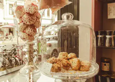 Eine Bäckereiauslage mit Mandelgebäck unter einer Glaskuppel und rosa bestreuten Baisers in einem hohen Glasgefäß, auf einem Fenstersims mit Blick auf die Straße draußen.