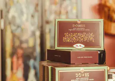 Ein Stapel grüner und brauner Schachteln mit der Aufschrift "Dômes aux Amandes Enrobés Chocolat Noir" steht in einem Regal, dahinter ein verschwommener, bunter Hintergrund.