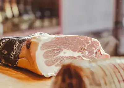 Nahaufnahme eines großen, teilweise eingewickelten Stücks Rohschinken mit marmoriertem Fett auf einer hölzernen Unterlage in einem Feinkostladen oder einer Metzgerei.