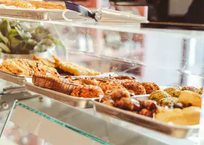 Eine Vitrine mit verschiedenen Backwaren und Gebäck in Metallschalen, mit einigen Blattpflanzen im Hintergrund und einer Nahaufnahme der Leckereien unter einem Glasregal.