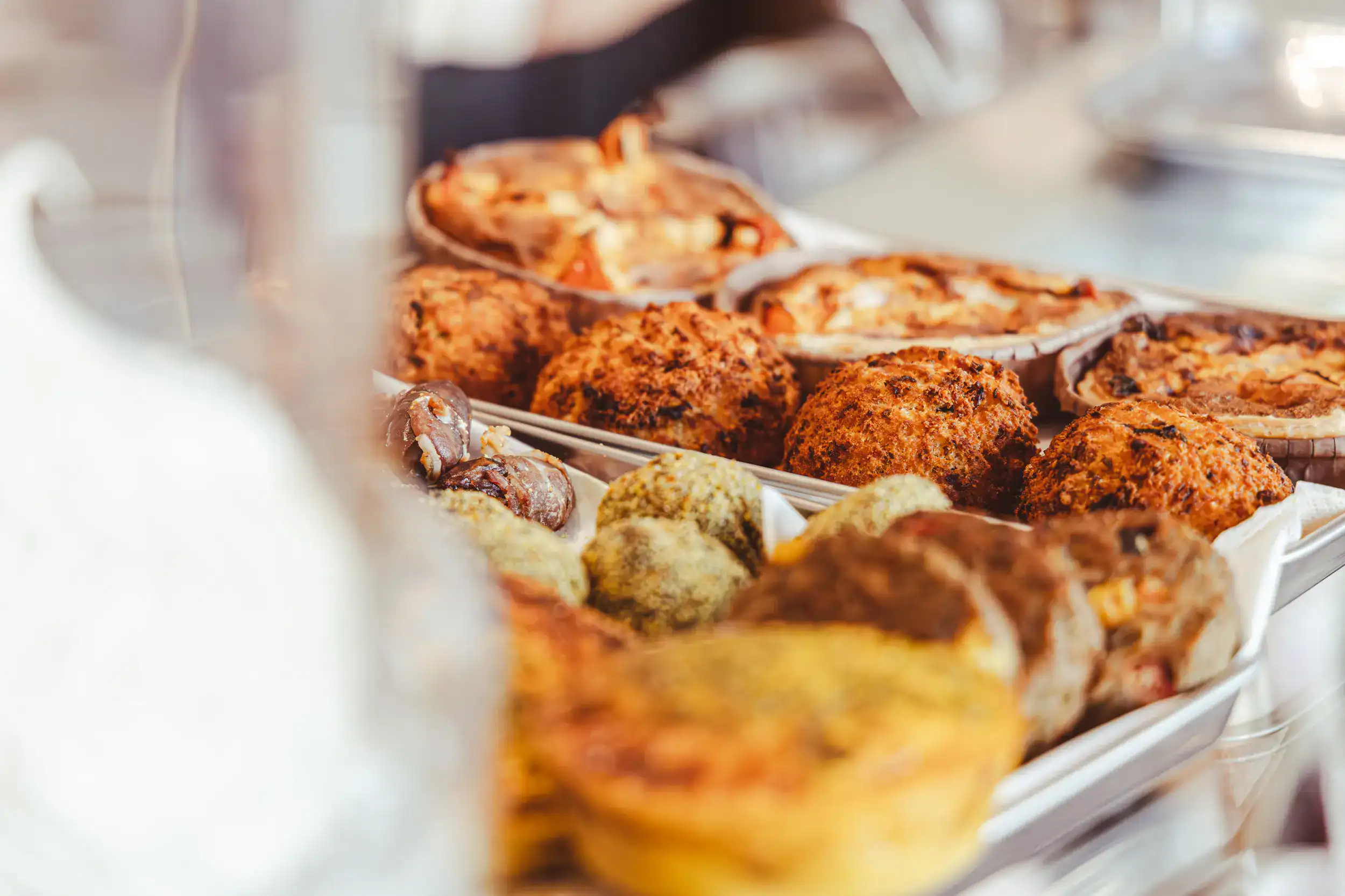 Eine Nahaufnahme von verschiedenen Backwaren, darunter Muffins, Kuchen und Gebäck, die auf Tabletts in einer Bäckerei ausgestellt sind. Die Leckereien sind goldbraun und sehen frisch gebacken aus.