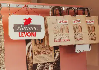 Auf einem Metallgestell sind Papiertüten mit dem Levoni-Branding und italienischen Delikatessenabbildungen zu sehen, daneben hängt ein Schild mit der Aufschrift stazione Levoni und einem roten Schweinelogo.