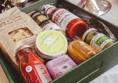 Ein grüner Geschenkkarton, gefüllt mit verschiedenen Gourmet-Lebensmitteln, darunter Nudeln, Nudelsaucen, Kräutersalz, Pesto, Olivenöl, Honig sowie Gläser und Dosen mit verschiedenen Gewürzen, die alle fein säuberlich im Inneren angeordnet sind.