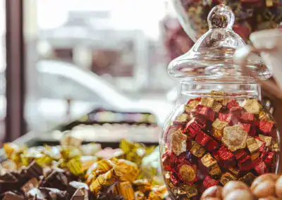 Ein Glas mit verpackten Pralinen in Gold, Rot und Grün steht auf einer Theke in einer Konditorei, umgeben von weiteren Süßigkeiten, mit einem Fenster und einer unscharfen Straßenszene im Hintergrund.