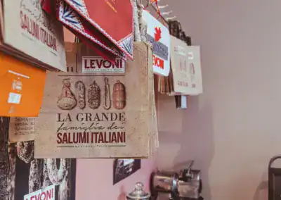 Mehrere dekorative Papiertüten hängen in einer Reihe an einer Wand, wobei die auffälligste Tüte die Aufschrift LA GRANDE famiglia dei SALUMI ITALIANI und Bilder verschiedener italienischer Wurstwaren trägt. Im Hintergrund sind weitere Markentüten zu sehen.