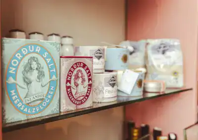 In einem Regal stehen verschiedene Dosen und Verpackungen von Nordur Meersalz, darunter blaue und violette Behälter mit Illustrationen, neben anderen Gewürzprodukten in pastellfarbenen Verpackungen.