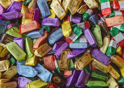 Ein großes Sortiment eingewickelter Bonbons in verschiedenen leuchtenden Farben, darunter Lila, Gold, Grün, Rosa und Blau, dicht an dicht gestapelt.