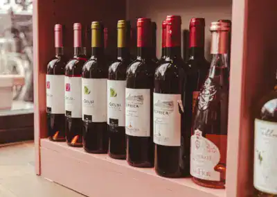 Auf einem rosafarbenen Holzregal in der Nähe eines Fensters ist eine Reihe von Weinflaschen mit verschiedenen Etiketten und Verschlussfarben aufgereiht, rechts daneben eine teilweise sichtbare Flasche Rosé.