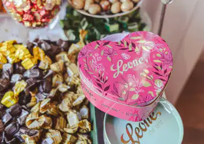 Eine rosafarbene, herzförmige Leone-Dose mit goldenen Blumenmustern steht zwischen verpackten Bonbons und anderen Leone-Dosen auf einem Tisch.