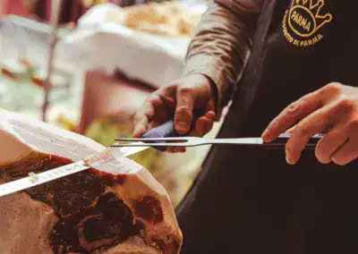 Eine Person in einer Parmaschürze schneidet den Schinken mit einem langen Messer und einer Gabel in dünne Stücke von einer großen Prosciutto-Keule.
