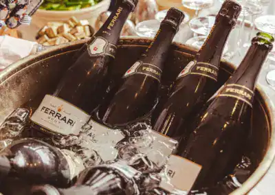 Fünf Flaschen Ferrari-Sekt kühlen in einem metallenen Eiskübel, umgeben von Eiswürfeln, während im Hintergrund mehrere leere Weingläser zu sehen sind.