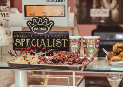 An der Theke eines Feinkostladens stehen ein Schild "Parma Prosciutto di Parma Specialist", ein Tablett mit aufgespießten Fleischproben, zwei gemusterte Pappbecher und einige Backwaren neben einer digitalen Waage.