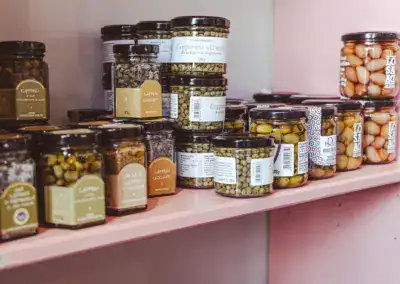 Gläser mit Kapern, Oliven und eingemachtem Gemüse stehen fein säuberlich auf rosa Regalen in einem Geschäft oder einer Speisekammer. Die Gläser haben verschiedene Etiketten und sind in Reihen gestapelt.