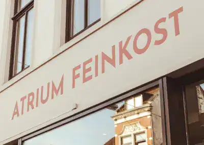 Auf einem Schaufensterschild steht in großen rosafarbenen Buchstaben ATRIUM FEINKOST über den Fenstern, die ein benachbartes Gebäude widerspiegeln.