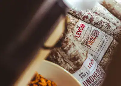 Nahaufnahme mehrerer eingewickelter italienischer Salamistangen mit der Aufschrift LEVON, die neben einer weißen Schale mit kleinen, knusprigen Snacks gestapelt sind. Das Bild hat eine warme, rustikale Ausstrahlung.