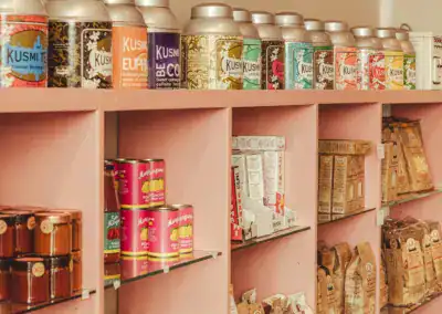 Eine Auslage mit farbenfrohen Dosen von Kusmi-Tee und verschiedenen verpackten Lebensmitteln, die fein säuberlich auf rosa Regalen in einem Geschäft angeordnet sind.