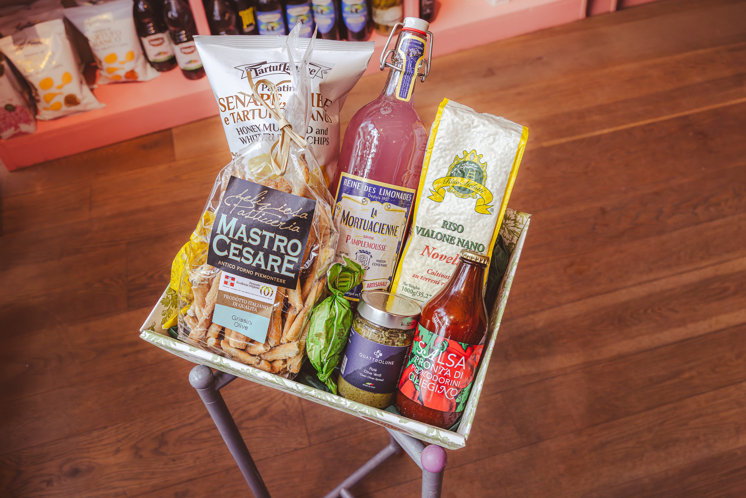 ATRIUM_Praesentkorb_2 Geschenkkorb mit italienischen Lebensmitteln wie Pasta, Grissini, Getränken und Käse auf einem Ständer in einem Geschäft mit Holzboden.
