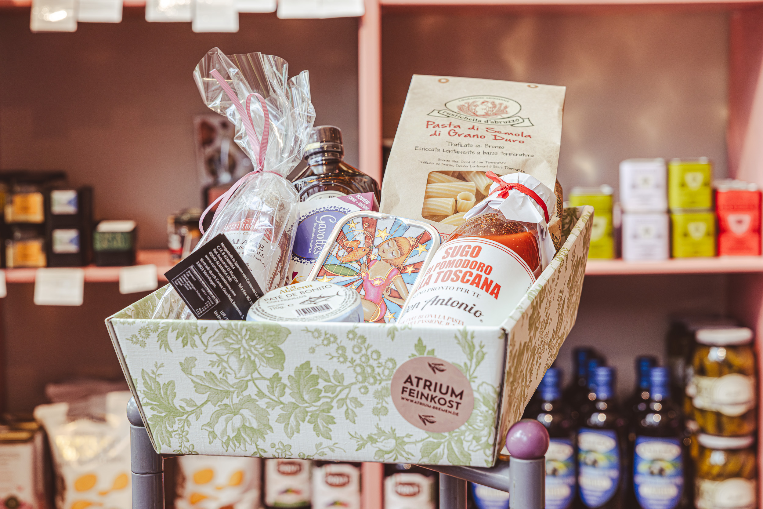 ATRIUM_Praesentkorb_3 Geschenkkarton mit Pasta, Tomatensauce, Olivenöl und Süßwaren im ATRIUM Feinkost in Bremen vor weiteren Geschenkkörben und Regalen.